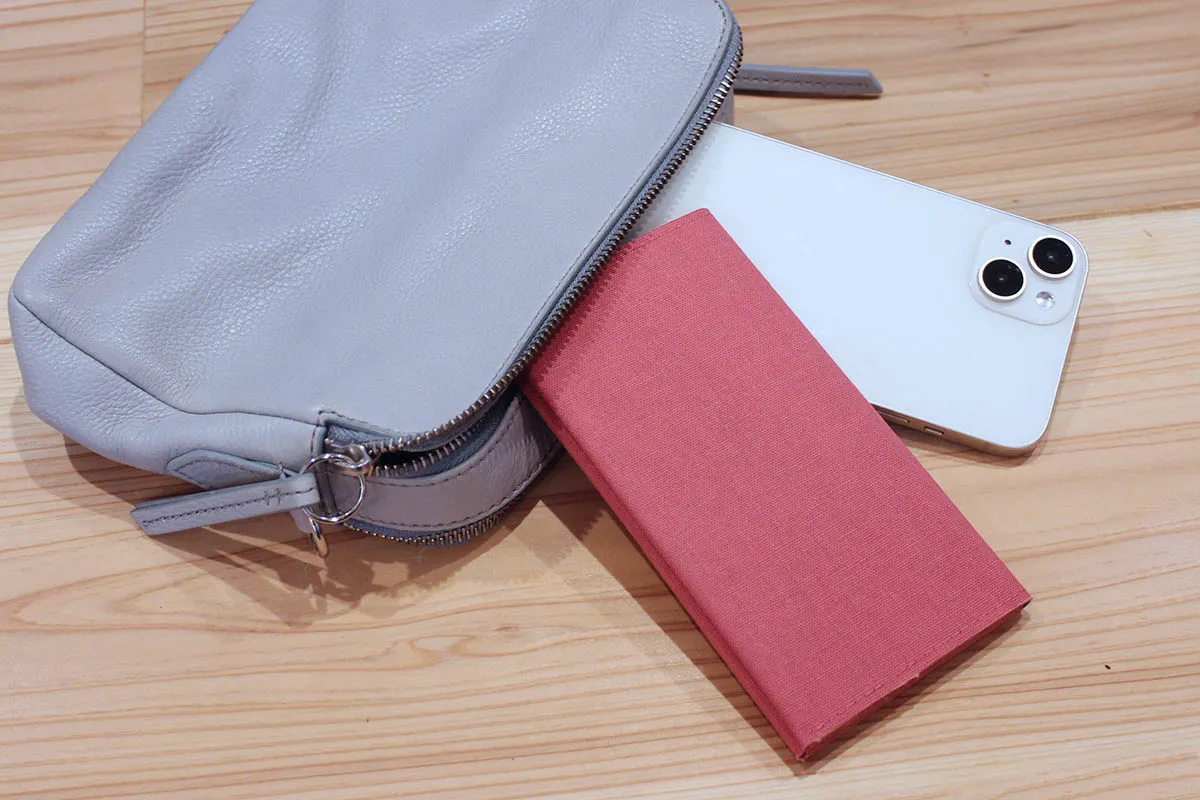 グレーのレザー製バッグから飛び出した白いスマートフォンと赤い手帳が、木製のテーブルの上に置かれている様子。